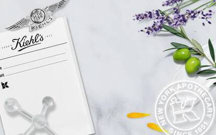 Alt-tekst: "Billede af Kiehl's branding med en historisk sort-hvid foto af apoteket, lavendel, oliven og blomster på en marmor-overflade."