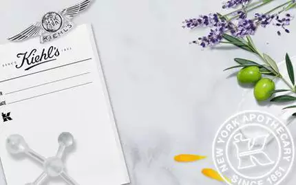 Alt-tekst: "Billede af Kiehl's branding med en historisk sort-hvid foto af apoteket, lavendel, oliven og blomster på en marmor-overflade."