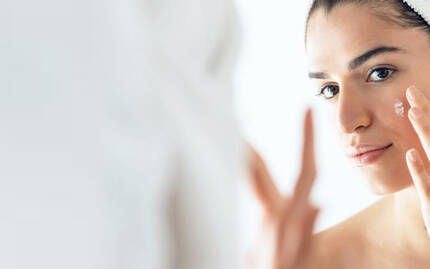 En ung kvinde med håndklaede om håret, der bruger ansigtscreme foran et spejl.