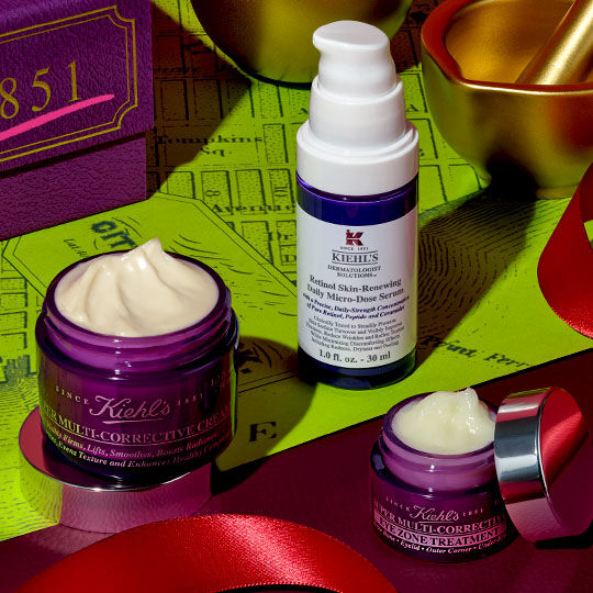 Tre Kiehl's Super Multi-Corrective produkter – en cremekrukke, en øjencreme og en serumflaske – er arrangeret på et rødt bånd, ved siden af en lilla aeske og guld julepynt, mod en limegrøn baggrund med et kortmønster.