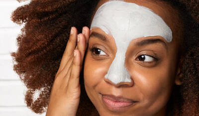 A young woman with curly hair applies a facial mask to her forehead, smiling softly.