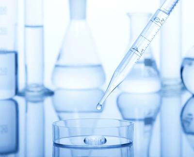 A dropper is poised above a petri dish with a single drop of liquid about to fall, surrounded by various laboratory glassware reflecting a blue hue.