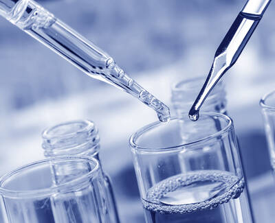 Two pipettes are positioned above a glass container filled with a clear liquid, with droplets poised to be released. The image has a blue-toned filter.