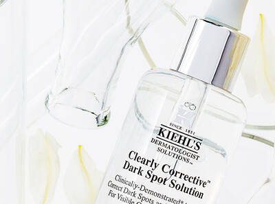 Kiehl's Clearly Corrective Dark Spot Solution in a glass bottle, surrounded by white petals and a transparent glass for a fresh look.