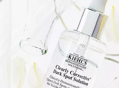 Kiehl's Clearly Corrective Dark Spot Solution in a glass bottle, surrounded by white petals and a transparent glass for a fresh look.