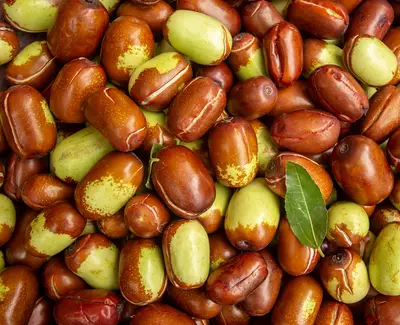 A close-up of a pile of jujube fruits, displaying a mix of red and green colors, some with a shiny appearance. A single green leaf is included among the fruits.