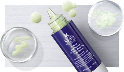 Alt text: A bottle of Kiehl's wrinkle-reducing serum is displayed with its product texture, alongside two small glass dishes containing a green gel and a white powder. The background is textured silver.