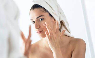 A woman with a towel on her head applies skincare cream to her face while looking in a mirror.