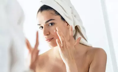 A woman with a towel on her head applies skincare cream to her face while looking in a mirror.