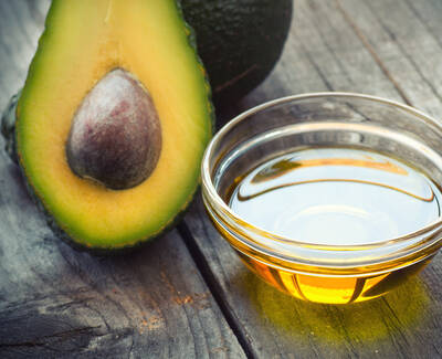 A sliced avocado revealing its seed next to a small glass bowl of golden avocado oil, all set against a rustic wooden background.
