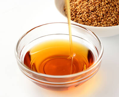 A glass bowl filled with golden brown oil, with a stream of oil being poured from above; a bowl of sesame seeds is in the background.