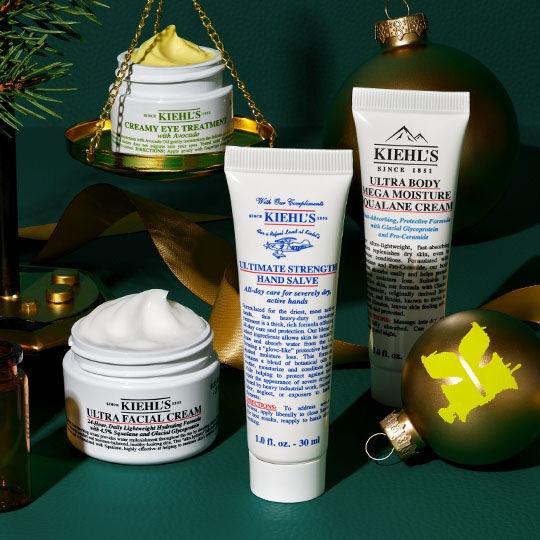 Et festligt arrangement af Kiehl's produkter, herunder Creamy Eye Treatment på gyldne skaller, Ultra Facial Cream, Ultimate Strength Hand Salve og Ultra Body Mega Moisture Squalane Cream, omgivet af grønne ornamenter, grangrene og guldbånd på en mørkegrøn baggrund.