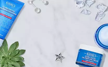 Alt-tekst: En flad laegning af Kiehl's hudplejeprodukter, herunder en ansigtsrens, toner og maske, omkranset af isterninger, en citron og grønne planter på en marmoroverflade.