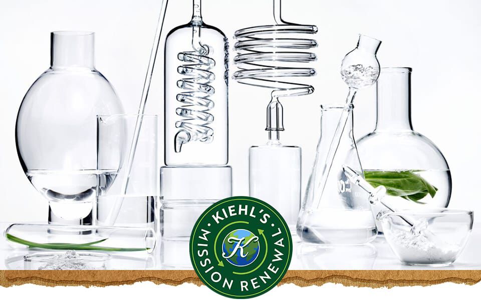 En samling af laboratoriekar af klart glas fyldt med vand og et grønt blad, med Kiehl's logo til højre.