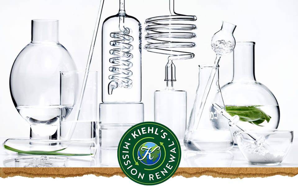 En samling af laboratoriekar af klart glas fyldt med vand og et grønt blad, med Kiehl's logo til højre.