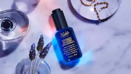 A blue bottle of Kiehl's Midnight Recovery Concentrate displayed on a marble surface, surrounded by lavender flowers and gold jewelry, with soft lighting creating a calming atmosphere.