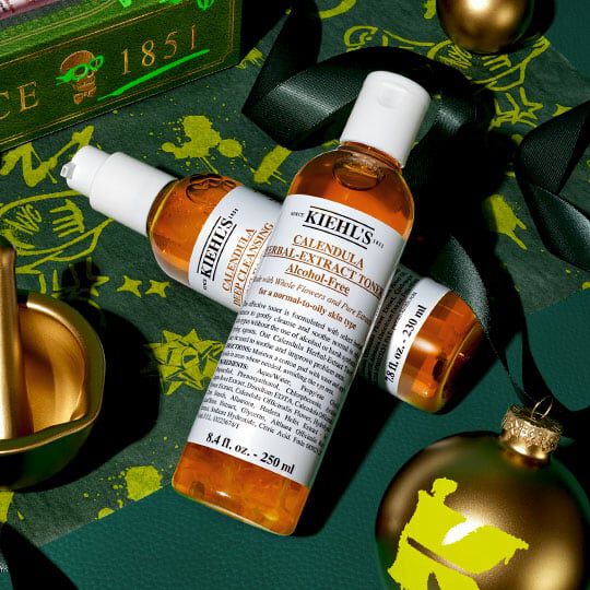 Kiehl's Calendula Herbal-Extract Toner og serum arrangeret med guldfarvede julepynt og grønt bånd på en festlig mørkegrøn baggrund.