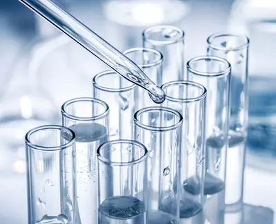 A close-up of clear glass test tubes filled with liquid, with a dropper dispensing a droplet into one of the tubes. The background is softly blurred, featuring laboratory equipment.