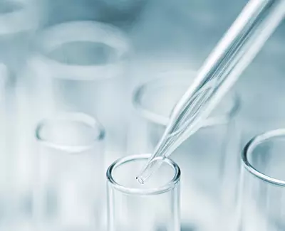 A glass pipette is poised above a test tube, preparing to transfer a liquid. The background features several blurred test tubes, creating a laboratory setting.