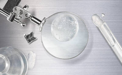 A close-up of a magnifying glass surrounded by laboratory equipment, including a test tube and a dropper, with droplets of liquid on a metallic surface.