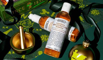 En grafik på en festlig mørkegrøn baggrund med guldaccenter viser to flasker: "Kiehl's Calendula Deep Cleansing Foaming Face Wash" og "Kiehl's Calendula Herbal-Extract Toner", begge med klar emballage og ravfarvet vaeske. Scenen inkluderer en guldmorter og -stamper, et guldornament med et grønt "K"-logo, sort bånd og baggrunden dekoreret med guldillustrationer.