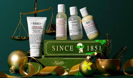 A festive flat lay image set against a dark green background showcases a selection of Kiehl's products amidst holiday-themed props. On the left, a golden balance scale holds what appears to be pinecones in its left pan. In the foreground, from left to right, are a white tube of Kiehl's Amino Acid Conditioner, a bottle of Kiehl's Bath and Shower Liquid Body Cleanser in "Grapefruit," a bottle of Kiehl's Amino Acid Shampoo, and a bottle of Kiehl's Creme de Corps. These products are arranged on and around two stacked dark green books. The top book features "SINCE 1851" in gold lettering with a skull graphic in green glasses, while the lower book shows "Kiehl's" also in gold. A golden ribbon weaves across the foreground from left to right. On the far right, a golden mortar and pestle contain a small green pine branch. Two ornaments are also visible: one is a golden bauble with a green 'K' logo, and another, partially visible, is a plain golden bauble