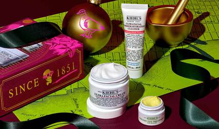 En festlig grafik, der viser en hvid tube med "KIEHL'S ULTRA FACIAL BARRIER-HYDRATING CLEANSER", en hvid krukke med "KIEHL'S ULTRA FACIAL CREAM" med synlig hvid creme og en lille hvid krukke med "KIEHL'S CREAMY EYE TREATMENT WITH AVOCADO" med synlig lysegul creme, alt sammen arrangeret på en mørkerød og grøn baggrund med et kortmønster. En guldmorter og -stamper er synlig øverst til højre. Øverst til venstre ses et guldjuleornament med et pink fugledesign og en rød gaveaeske med "SINCE 1851" og et dødningehoveddesign. Sort bånd er draperet omkring produkterne.