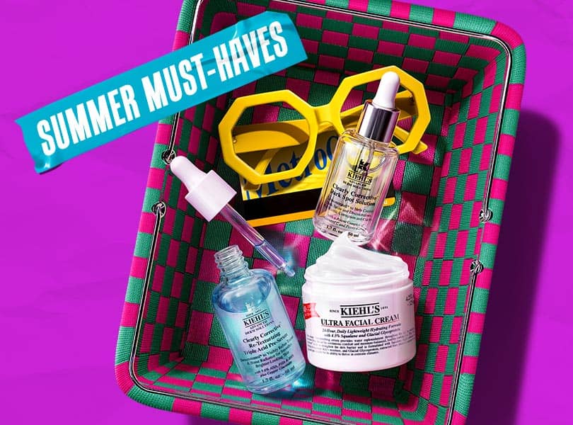 A green and pink woven basket containing Kiehl's skincare products including Ultra Facial Cream and serums, with yellow sunglasses and a 'Summer Must-Haves' sticker.